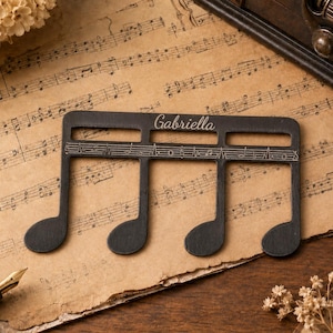 Puede incluir: Un objeto negro y plano con forma de nota musical que presenta el nombre Gabriella grabado en la parte superior y un pentagrama horizontal con notas grabadas en el centro. El art&iacute;culo tiene un acabado mate liso y cuatro plicas.