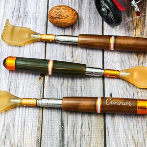 Puede incluir: Tres rascadores de espalda de madera con un extremo rascador en forma de mano. Los rascadores tienen detalles en marrón, plateado y naranja. Un rascador de espalda está grabado con el nombre "Connor". Los artículos están sobre una superficie de madera.