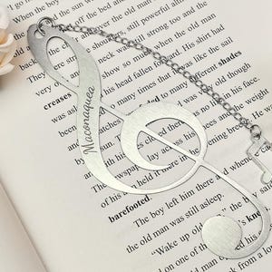 May include: A silver-colored metal bookmark in the shape of a treble clef, with a chain and a small musical note charm. The word "Maconaquea" is engraved on the bookmark. The bookmark rests on the pages of a book.