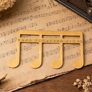 Puede incluir: Un marcap&aacute;ginas de metal dorado con forma de semicorchea y una pauta horizontal recortada en el centro. La pauta presenta notas musicales y s&iacute;mbolos grabados, incluyendo una clave de sol, negras y un signo de bemol.
