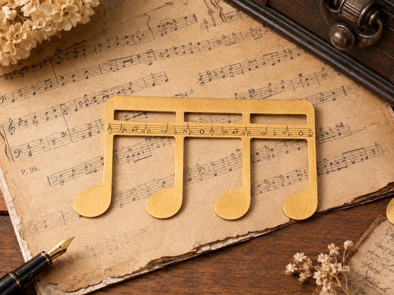Puede incluir: Un marcap&aacute;ginas de metal dorado con forma de semicorchea y una pauta horizontal recortada en el centro. La pauta presenta notas musicales y s&iacute;mbolos grabados, incluyendo una clave de sol, negras y un signo de bemol.
