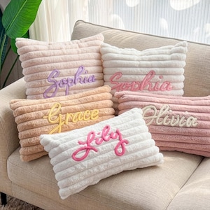 Puede incluir: Un conjunto de cinco cojines de pana en varios colores, incluyendo blanco, rosa y beige. Cada cojín presenta un nombre bordado: Sophia, Grace, Olivia y Lily. Los cojines están colocados sobre un sofá beige.