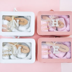 Może przedstawiać: Cztery różowe i białe pudełka prezentowe z przezroczystymi pokrywkami. Każde pudełko zawiera szczotkę do włosów, dwie spinki do włosów, opaskę na głowę i drewniany znaczek z imieniem ozdobiony kamieniami szlachetnymi. Imię to Nicole, Hailey, Megan i Emma.