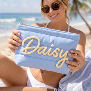 Puede incluir: Un bolso de mano acolchado azul claro con cremallera dorada. La palabra "Daisy" está bordada en hilo amarillo en la parte delantera. El bolso se muestra sobre un fondo de playa.