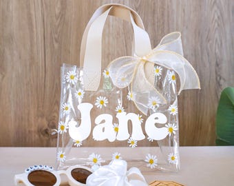 May include: Clear plastic tote bag with a white daisy print and the name "Jane" in white letters. The bag has a cream-colored handle and a cream-colored bow.