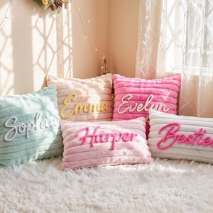 May include: A collection of five plush, ribbed-texture throw pillows in pastel shades of mint green, light pink, hot pink, and white. Each pillow features a raised, cursive name or word appliqué in a contrasting color, reading Sophia, Emma, Evelyn, Harper, and Besties.