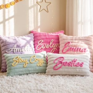 May include: A collection of five plush, horizontally ribbed throw pillows in pastel colors including lavender, hot pink, light pink, sage green, and white. Each pillow features raised, cursive lettering reading Amelia, Evelyn, Emma, Stay Cozy, and Besties.