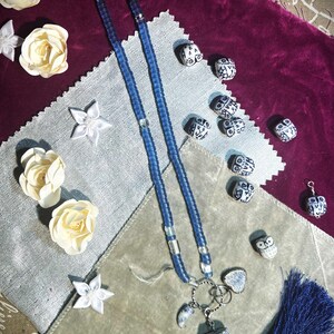 May include: A blue beaded necklace with a tassel and stone pendants. The necklace features a silver clasp and is adorned with small owl-shaped beads. Decorative white flowers and fabric pieces surround the necklace.