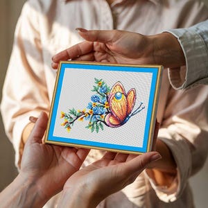 Puede incluir: Una obra de arte en punto de cruz enmarcada con un diseño de mariposa y flores. La mariposa tiene tonos naranja, amarillo y morado, con una gema azul. Las flores son azules y amarillas, sobre un fondo blanco con borde azul y marco dorado.