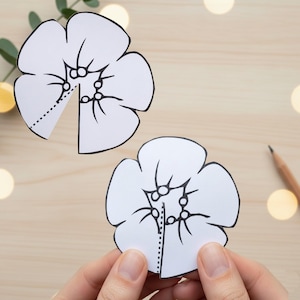 May include: Two white paper flower cutouts with black outlines and dotted lines, one held by hands. The flowers have detailed petal and center designs. A pencil and greenery are in the background on a light wood surface.