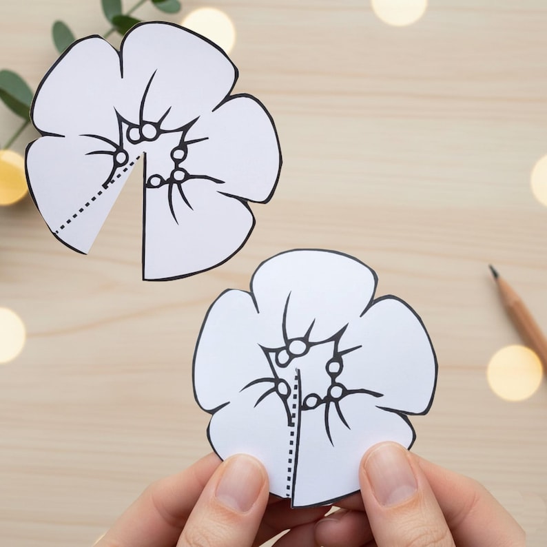 May include: Two white paper flower cutouts with black outlines and dotted lines, one held by hands. The flowers have detailed petal and center designs. A pencil and greenery are in the background on a light wood surface.