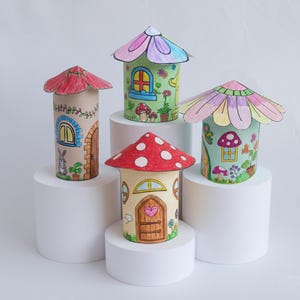 Puede incluir: Cuatro casas de hadas hechas a mano. Cada casa está diseñada de forma única con detalles coloridos, incluyendo acentos de hongos y flores, y una variedad de estilos de techo. Las casas se exhiben sobre pedestales cilíndricos blancos.