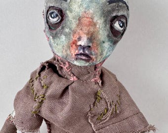 May include: A handmade doll with a pale green face, dark eyes, and a distressed appearance. The doll wears a brown fabric outfit with pink and green stitching details. The doll's head has a textured surface.