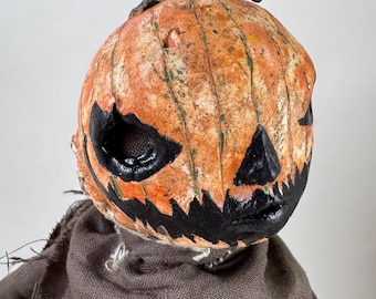 May include: A Halloween-themed decorative figure with a pumpkin head. The pumpkin is painted orange with black facial features and a dark green stem. It is attached to a brown fabric body, creating a spooky, handcrafted aesthetic.