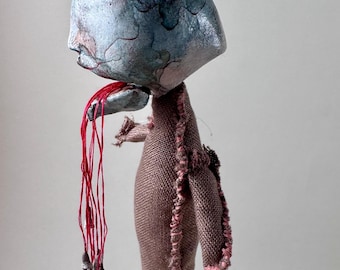 May include: A handmade art doll with a blue and gray head, a fabric body, and red string hair. The doll has a unique, textured appearance and is suspended by a red string. The doll's body is a muted pink color with frayed edges.