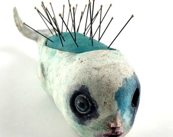 May include: A ceramic pincushion shaped like a fish with a blue and white painted design. The fish has large eyes and a small mouth. The pincushion is covered in black pins with gold heads.