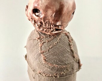 May include: A small, ceramic doll with a stitched-on fabric body. The doll has a grotesque, human-like face with a large, open mouth and a single, dark eye.