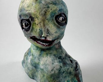 May include: A handmade sculpture of a humanoid figure with a large head and a small body. The figure is painted in shades of green, blue, and white, with dark eyes and a red mouth. The sculpture has a slightly unsettling appearance.