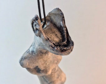 May include: A white ceramic pendant shaped like a human torso with a textured surface. The pendant is hanging from a black cord.