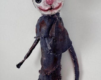 May include: A handmade doll with a pale, textured, round head featuring large black eye sockets, a pink nose, and a red painted mouth. The body is constructed from dark blue denim fabric with frayed edges, visible stitching, and dark brown stained patches.