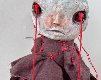 May include: A handmade doll with a pale, textured head and red yarn flowing from the eye sockets. The doll wears a dark brown fabric outfit with red stitching. The doll's face has a somber expression.