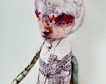 May include: A handmade doll with a pale, textured, rounded head featuring eyes stitched shut with bright red thread. The torso is covered in off-white fabric printed with a moth illustration and the text Noctua (Ophideres).