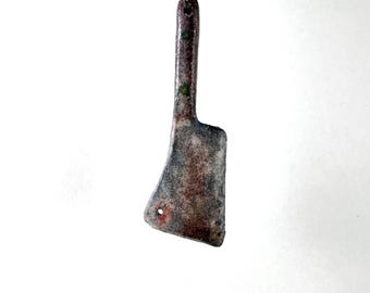 May include: A rustic cleaver pendant hangs from a silver ball chain. The pendant features a weathered, textured appearance with shades of brown, gray, and green. The cleaver design includes a small hole near the blade's edge, adding to its unique charm.