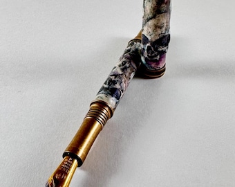 May include: A fountain pen featuring a brass grip section with ribbed detailing and a gold-toned nib. The barrel and cap are crafted from a marbled stone material displaying shades of grey, white, and purple. The cap features a brass top band.