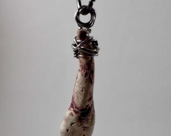 May include: A pendant with a unique design, featuring a light-colored, elongated object with red markings, suspended from a silver-colored chain. The object is wrapped with silver wire. The background is a plain, light gray.
