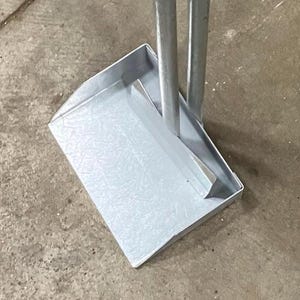 May include: A silver metal dustpan with a long handle. The dustpan is rectangular with a slightly textured surface. Two silver poles are attached to the dustpan, forming the handle.