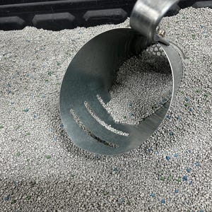 May include: A metal scoop with holes is partially submerged in a container of grey cat litter. The litter is made up of small, round pellets.