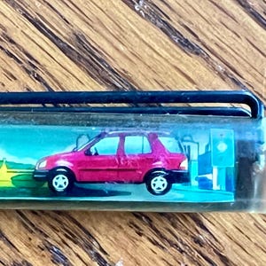 May include: A clear plastic pen barrel containing a miniature graphic insert featuring a bright magenta SUV parked near a yellow bridge and a green landscape. The pen has a black plastic clip attached to the top of the transparent cylindrical casing.
