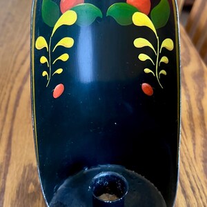 May include: Black metal candle holder with a decorative floral design in red, yellow, and green. The candle holder has a rounded top and a small circular base for the candle. The edges are trimmed in gold.