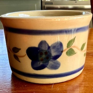 May include: A ceramic container with a beige background, blue stripes, and a blue floral design. Green leaves and brown vines are painted around the flower. The container is cylindrical with a slightly flared rim.