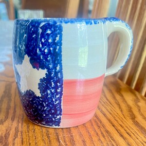 May include: A ceramic mug featuring a hand-painted design of the Texas state flag. The mug displays a vertical blue stripe with a white star on the left side, and horizontal white and red stripes on the right side, with a curved handle on the right.