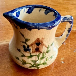 May include: A small, ceramic pitcher with a cream-colored body and a blue rim and handle. The pitcher is decorated with a brown birdhouse design and green foliage. The pitcher has a small spout for pouring.