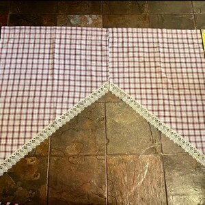 May include: A pair of plaid curtains with a red and white pattern, trimmed with a cream-colored lace. A yellow measuring tape is placed along the edge of the curtain, indicating its length.