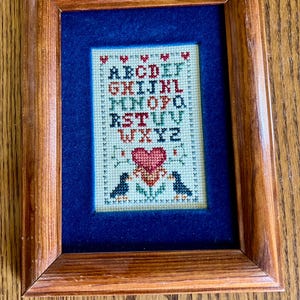 May include: Framed cross-stitch sampler with a blue background. The sampler features the alphabet in red and green, a red heart, and two birds. The frame is made of wood.