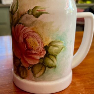 May include: White ceramic mug with a large, hand-painted red rose and green leaves. The mug has a handle and a wide base. The background has soft green and peach tones.