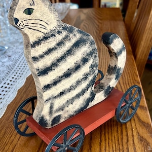 May include: A vintage wooden cat figurine with black and cream stripes, green eyes, and a black nose. The cat sits on a red wooden cart with black spoked wheels. The cat's tail curves upwards.
