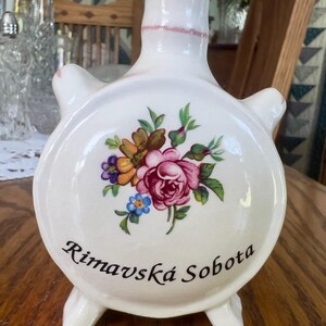 May include: A white ceramic flask with a rounded body, a narrow neck, and two small side handles. The front features a colorful floral bouquet of pink, yellow, and blue flowers, with the black text Rimavská Sobota printed below the design.