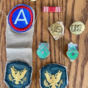 May include: A collection of vintage military insignia and patches. Includes a blue and red patch with a white "A", a red and white ribbon, two "US" pins, two green and silver pins, and patches with gold eagles.