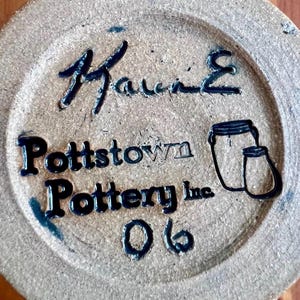 May include: A circular tan ceramic base featuring a black handwritten signature at the top, the text Pottstown Pottery Inc. in the center, a line drawing of two jars on the right, and the number 06 printed at the bottom of the piece.