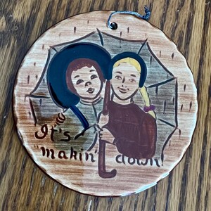 May include: Round, ceramic ornament with a painted illustration of two children under an umbrella. The children are wearing hats and holding an umbrella. The text "It's makin' down!" is written on the ornament. A blue string is attached for hanging.