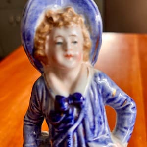 May include: A porcelain figurine of a young person wearing a blue hat and matching jacket with a bow. The figurine has a painted face with rosy cheeks and curly blonde hair. The jacket has gold accents.