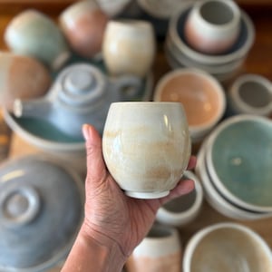 Puede incluir: Un vaso de cerámica blanco roto hecho a mano con forma redondeada, sostenido en una mano. El vaso tiene un sutil degradado terroso de colores marrón y crema. Otras piezas de cerámica, incluyendo cuencos y una tetera, son visibles en el fondo.