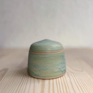 Puede incluir: Un pequeño tarro de cerámica con tapa, que presenta un esmalte moteado en verde claro y azul sobre una base lisa de color beige. El recipiente tiene un cuerpo cilíndrico y una tapa cónica ligeramente puntiaguda que encaja en el borde.