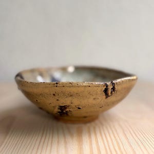 May include: A small, round ceramic bowl with a textured, mottled tan and brown exterior. The rim is uneven and slightly irregular, featuring dark brown speckles and patches of glaze that transition into a lighter, muted interior surface.