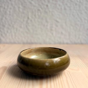 Op de afbeelding: Een kleine, ronde keramische kom met een glanzende afwerking. De buitenkant heeft een diepe olijfgroene glazuurlaag, terwijl de binnenkant overgaat in een lichtere, bleke saliegroene tint. De rand is glad en afgerond met een eenvoudig ontwerp.