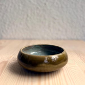 Puede incluir: Un pequeño cuenco redondo de cerámica con un exterior verde oliva brillante y un interior esmaltado en azul claro liso. El cuenco presenta una silueta curva de perfil bajo con un borde grueso, mostrando un contraste distintivo entre el tono terroso exterior y el color azul suave y apagado del interior.